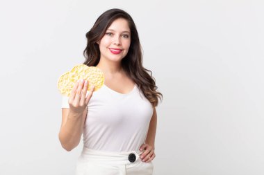 Güzel bir kadın mutlu bir şekilde gülümsüyor, eli kalçasında, kendinden emin ve elinde diyet pirinç keki tutuyor.