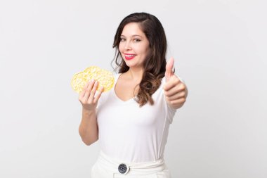 Güzel bir kadın gururlu, olumlu gülümsüyor başparmakları havada ve elinde diyet pirinç keki tutuyor.