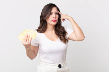 Güzel kadın şaşkın ve şaşkın hissediyor, deli olduğunu gösteriyor ve elinde diyet pirinç keki tutuyor.