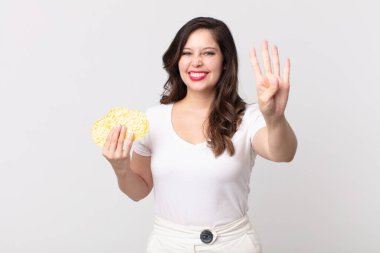 Güzel bir kadın gülümsüyor ve arkadaş canlısı görünüyor, dört numarayı gösteriyor ve elinde diyet pirinç keki tutuyor.