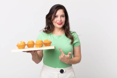 Güzel bir kadın neşeyle gülümsüyor, mutlu hissediyor ve bir tarafı işaret ediyor ve bir kek tepsisi tutuyor.