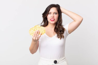 Güzel bir kadın stresli, endişeli ya da korkmuş, elleri başının üstünde ve elinde diyet pirinç keki tutuyor.