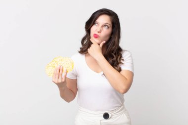 Güzel bir kadın düşünüyor, kuşkulu ve şaşkın hissediyor ve elinde diyet pirinç keki tutuyor.