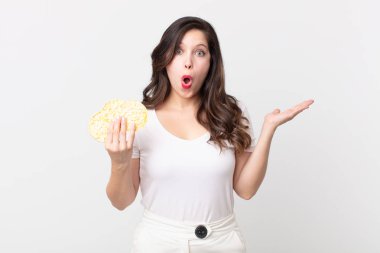 Güzel bir kadın şaşırmış ve şok olmuş görünüyor, çenesi düşmüş, elinde bir nesne ve elinde diyet pirinç keki tutuyor.