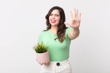 Güzel bir kadın gülümsüyor ve arkadaş canlısı görünüyor, beş numarayı gösteriyor ve elinde dekoratif bir bitki tutuyor.