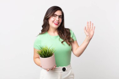 Güzel bir kadın mutlu bir şekilde gülümsüyor, el sallıyor, seni selamlıyor ve dekoratif bir bitki tutuyor.
