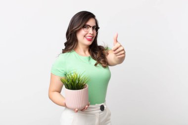 Güzel bir kadın gururlu, olumlu gülümsüyor başparmakları havada ve elinde dekoratif bir bitki tutuyor.