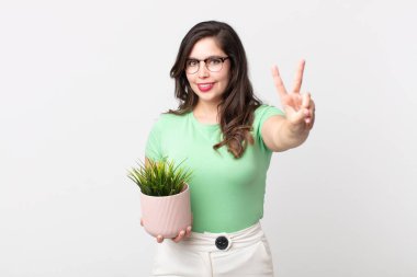 Güzel bir kadın gülümsüyor ve arkadaş canlısı görünüyor. İki numarayı gösteriyor ve elinde dekoratif bir bitki tutuyor.