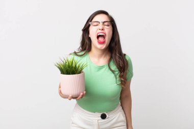 Güzel bir kadın agresif bir şekilde bağırıyor, kızgın görünüyor ve elinde dekoratif bir bitki tutuyor.