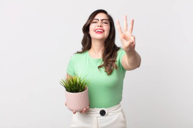 Güzel bir kadın gülümsüyor ve arkadaş canlısı görünüyor, üç numarayı gösteriyor ve elinde dekoratif bir bitki tutuyor.