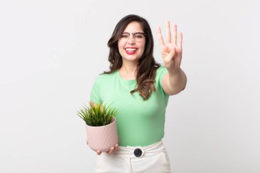 Güzel bir kadın gülümsüyor ve arkadaş canlısı görünüyor, dört numarayı gösteriyor ve elinde dekoratif bir bitki tutuyor.
