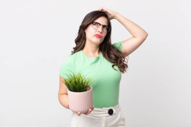 Güzel bir kadın şaşkın ve kafası karışmış hissediyor. Kafasını kaşıyor ve elinde dekoratif bir bitki tutuyor.
