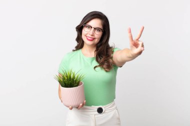 Güzel bir kadın gülümsüyor ve mutlu görünüyor, zafer ya da barış işareti yapıyor ve elinde dekoratif bir bitki tutuyor.