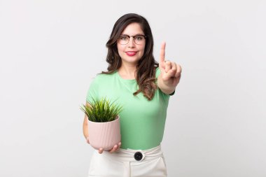 Güzel bir kadın gülümsüyor ve arkadaş canlısı görünüyor. Bir numarayı gösteriyor ve elinde dekoratif bir bitki tutuyor.