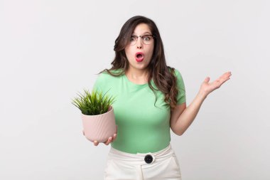 Güzel bir kadın şaşırmış ve şok olmuş görünüyor, çenesi düşmüş, elinde bir nesne, elinde dekoratif bir bitki tutuyor.