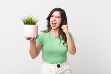 Güzel bir kadın kızgın bir ifadeyle saldırganca bağırıyor ve elinde dekoratif bir bitki tutuyor.