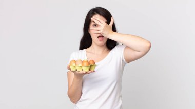 Güzel bir kadın şok olmuş, korkmuş ya da korkmuş görünüyor. Yüzü elle örtülmüş ve elinde bir yumurta kutusu tutuyor.