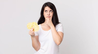 Güzel bir kadın düşünüyor, kuşkulu ve şaşkın hissediyor ve elinde diyet pirinç keki tutuyor.