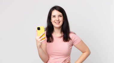 Güzel bir kadın mutlu bir şekilde gülümsüyor, eli kalçasında ve kendine güvenen akıllı bir telefon kullanıyor.