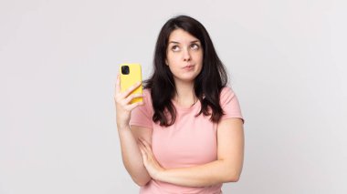 Güzel bir kadın omuz silkiyor, kafası karışık ve akıllı bir telefon kullanıyor.