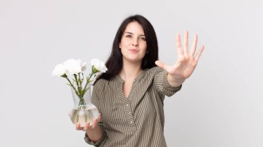 Güzel bir kadın gülümsüyor ve arkadaş canlısı görünüyor, beş numarayı gösteriyor ve elinde dekoratif bir çiçek tutuyor. Kulaklıklı bir yardımcı ajan.