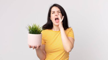 Güzel bir kadın kendini mutlu hissediyor, elleri ağızının yanında ve elinde dekoratif bir bitki tutuyor.