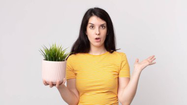 Güzel bir kadın şaşırmış ve şok olmuş görünüyor, çenesi düşmüş, elinde bir nesne, elinde dekoratif bir bitki tutuyor.