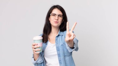 Güzel bir kadın gülümsüyor ve arkadaş canlısı görünüyor. İki numarayı gösteriyor ve elinde kahve tutuyor.