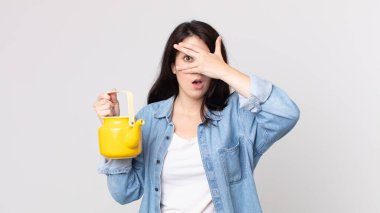 Güzel bir kadın şok olmuş, korkmuş ya da korkmuş görünüyor. Yüzü elle kaplanmış ve elinde bir çaydanlık tutuyor.