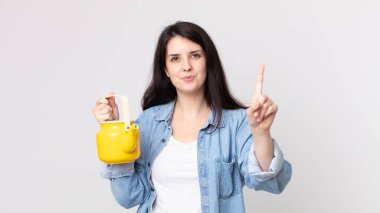 Güzel bir kadın gülümsüyor ve arkadaş canlısı görünüyor. Bir numarayı gösteriyor ve elinde çaydanlık tutuyor.