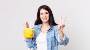 Güzel bir kadın gülümsüyor ve arkadaş canlısı görünüyor. İki numarayı gösteriyor ve elinde çaydanlık tutuyor.