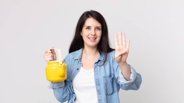 Güzel bir kadın gülümsüyor ve arkadaş canlısı görünüyor. Dört numarayı gösteriyor ve elinde çaydanlık var.