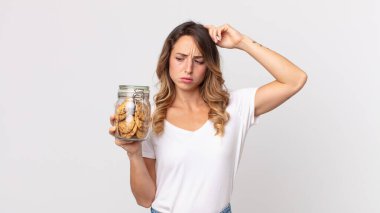 Kafası karışmış, kafası karışmış, kafasını kaşıyan ve elinde cam bir kurabiye şişesi tutan ince bir kadın.