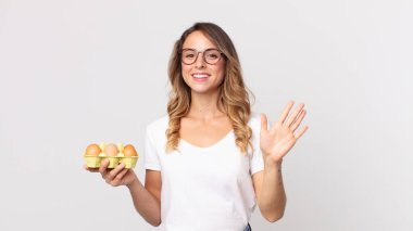 Güzel, ince bir kadın mutlu bir şekilde gülümsüyor, el sallıyor, seni selamlıyor ve bir yumurta kutusu tutuyor.