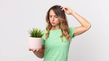 Kafası karışmış, kafası karışmış, başını kaşıyan ve elinde dekoratif bir bitki tutan ince bir kadın.
