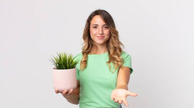 Oldukça zayıf bir kadın mutlu bir şekilde gülümsüyor ve bir konsept sunuyor ve elinde dekoratif bir bitki tutuyor.