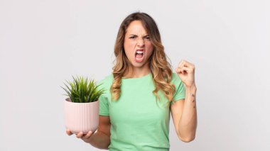 Oldukça zayıf bir kadın kızgın bir ifadeyle saldırganca bağırıyor ve elinde dekoratif bir bitki tutuyor.