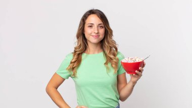 Oldukça zayıf bir kadın mutlu bir şekilde gülümsüyor, eli kalçasında, kendinden emin ve elinde bir kahvaltı kasesi tutuyor.