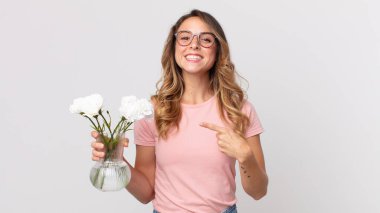 Oldukça zayıf bir kadın neşeli bir şekilde gülümsüyor, mutlu hissediyor ve yan tarafı işaret ediyor ve elinde dekoratif çiçekler tutuyor.