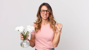 Oldukça zayıf bir kadın kızgın bir ifadeyle saldırganca bağırıyor ve elinde dekoratif çiçekler tutuyor.