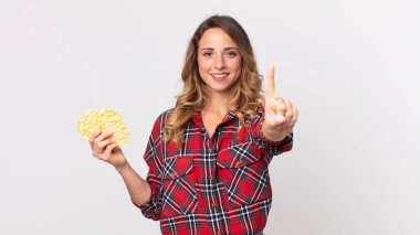 Çok zayıf bir kadın gülümsüyor ve arkadaş canlısı görünüyor. Bir numarayı gösteriyor ve elinde diyet pirinç keki tutuyor.