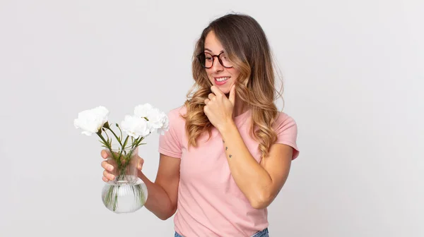 pretty thin woman smiling with a happy, confident expression with hand on chin and holding decorative flowers