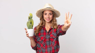 Oldukça zayıf bir kadın gülümsüyor ve arkadaş canlısı görünüyor, elinde kaktüs tutan üç numarayı gösteriyor. çiftçi kavramı