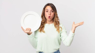 Oldukça zayıf bir kadın şaşırmış ve şok olmuş görünüyor, çenesi düşmüş, elinde bir nesne, elinde boş bir tabak, elinde boş bir tabak tutuyor.