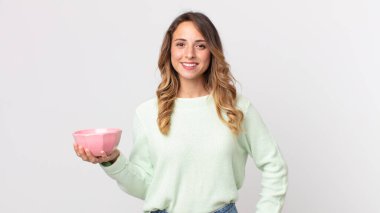 Oldukça zayıf bir kadın mutlu bir şekilde gülümsüyor, eli kalçasında, kendinden emin ve elinde boş bir tencere tutuyor.