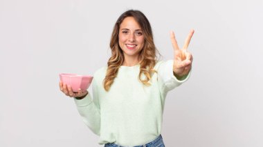 Çok zayıf bir kadın gülümsüyor ve arkadaş canlısı görünüyor. İki numarayı gösteriyor ve elinde boş bir tencere tutuyor.