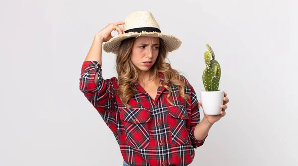 pretty thin woman feeling puzzled and confused, scratching head holding a cactus. farmer concept