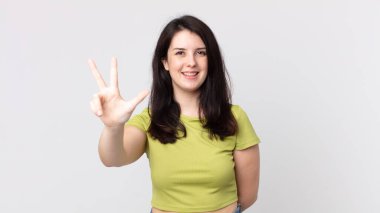 Güzel bir kadın gülümsüyor ve arkadaş canlısı görünüyor. Üç ya da üç numarayı gösteriyor. Elleri önde, geri sayıyor.