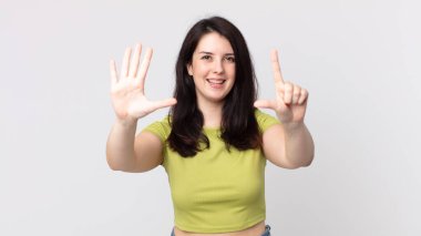 Güzel bir kadın gülümsüyor ve dost canlısı görünüyor, 7 ya da 7 numarayı gösteriyor, eli önde, geriye doğru sayıyor.