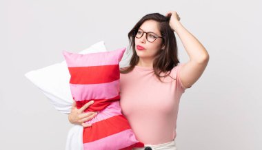 Güzel bir kadın şaşkın ve kafası karışmış hissediyor. Pijama giymiş ve elinde yastıkla kafasını kaşıyor.